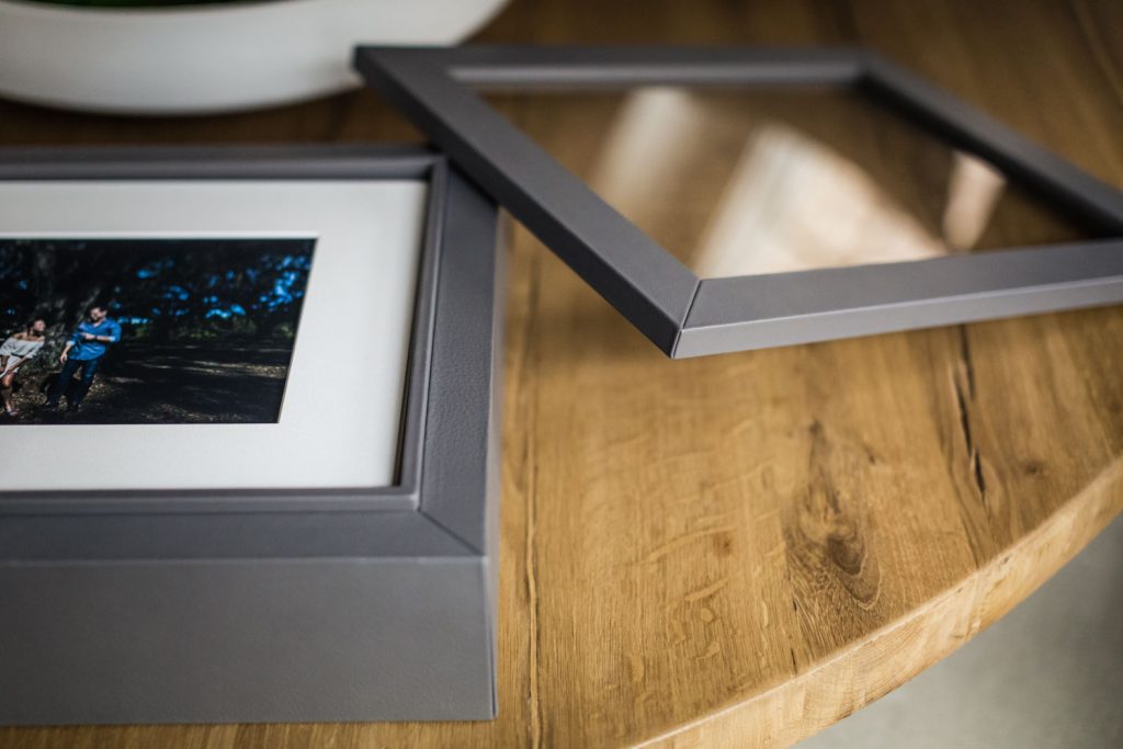 A gray picture frame with its glass cover removed sits on a wooden table. A photo is mounted inside the frame, partially visible, showing people outdoors under trees.