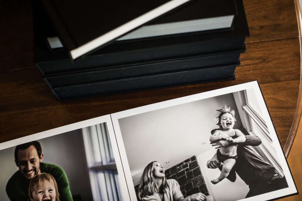 A stack of photo albums sits on a wooden table next to open pages showing black-and-white photos of smiling adults and a laughing baby being lifted into the air.