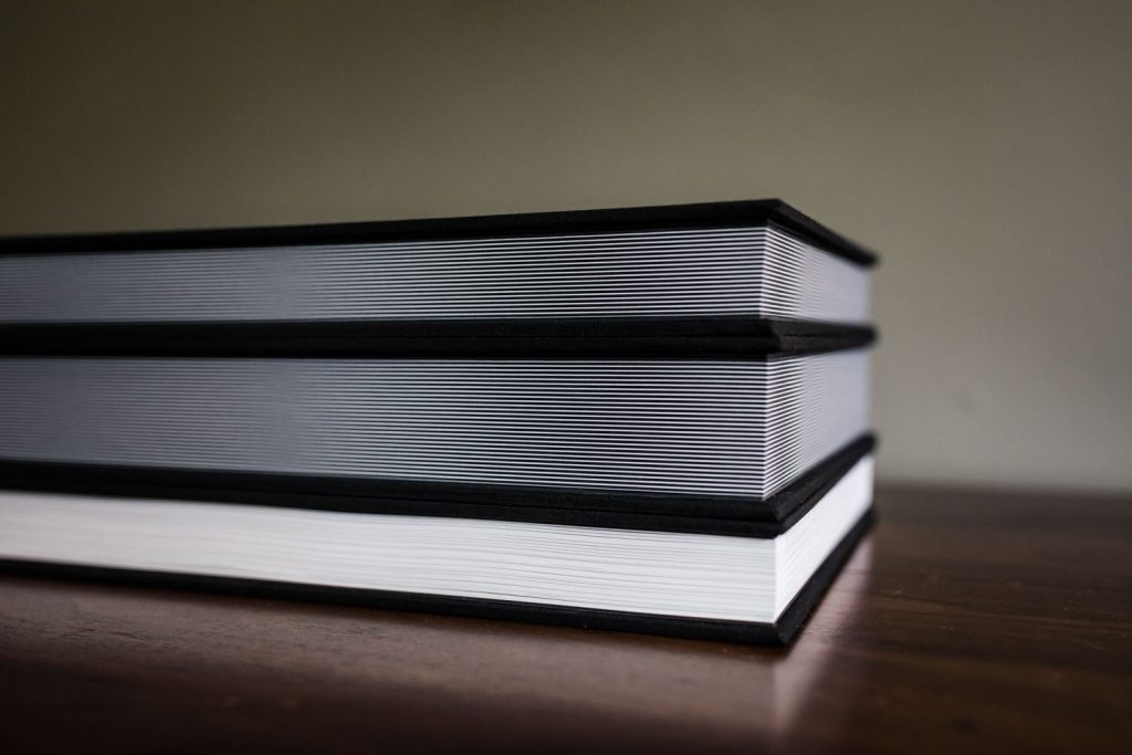 Two thick, hardcover books with black edges are stacked on a wooden surface, viewed from the side, showing the crisp, white pages and dark covers. The background is plain and out of focus.