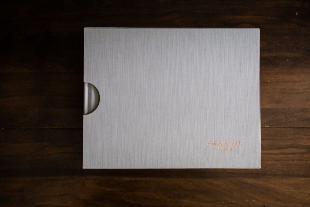 A gray textured box with a semicircular cutout on the side sits on a dark wooden surface. Orange text on the lower right corner reads Ashley & Teddy 10.13.18.