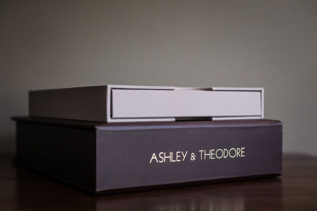 Two elegant boxes stacked on a wooden surface; the bottom box is dark with ASHLEY & THEODORE printed on the side, while the top box is light gray with a textured finish.