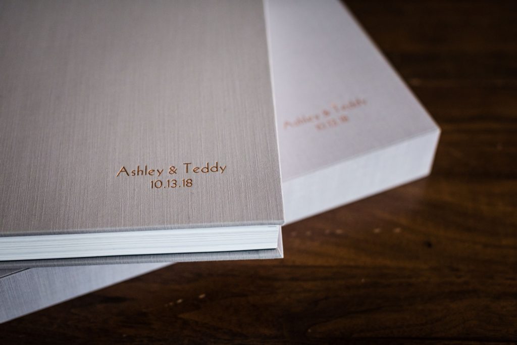 Close-up of two elegant photo albums with light gray textured covers, one on top of the other. The top album is embossed with Ashley & Teddy 10.13.18 in gold lettering. Both are resting on a wooden surface.