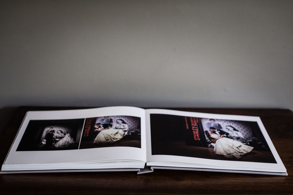 An open photo album lies on a dark wooden surface, displaying vintage-style wedding photos across two pages, featuring a bride in a white dress and a groom in a suit against a patterned backdrop.