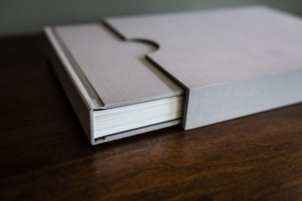 A close-up of a stack of photo prints or cards in a light gray, textured slipcase on a dark wooden surface. The slipcase is partially open, revealing the neatly stacked contents inside.