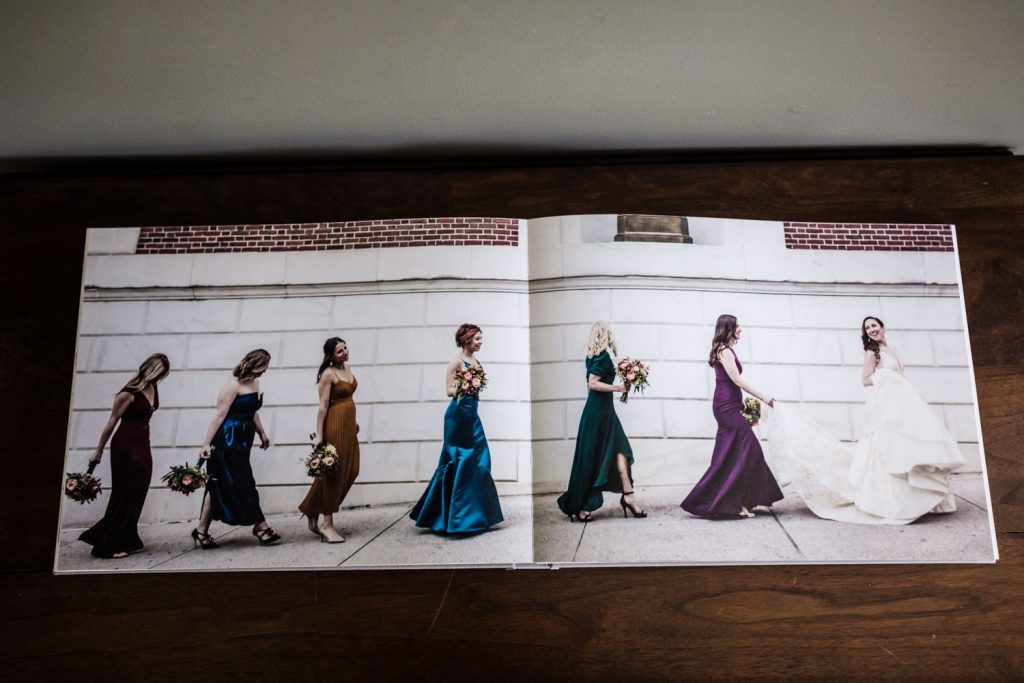 A photo album open to a spread showing five women in colorful dresses and one in a white wedding gown walking together outside along a stone wall, each holding a bouquet.