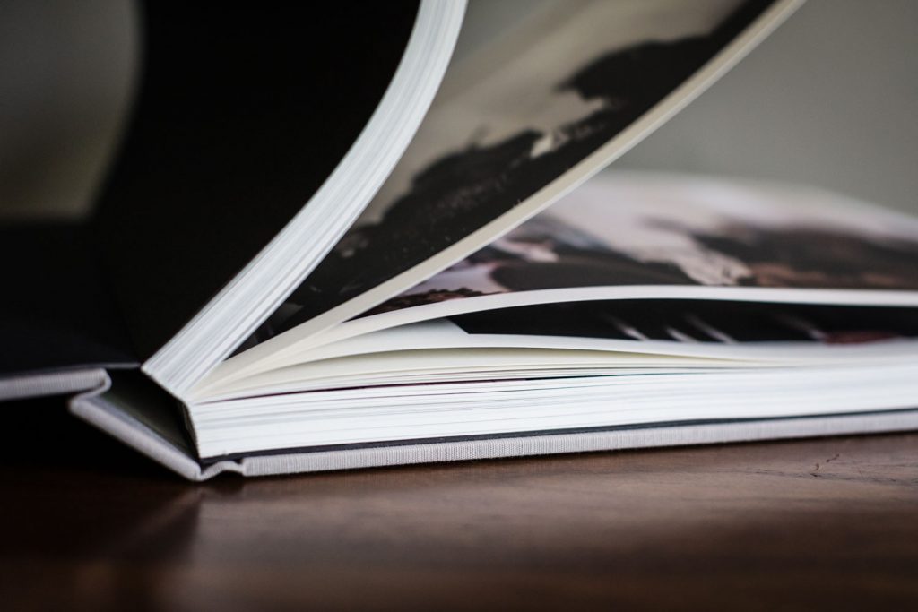 Close-up of an open photo book with thick pages, lying on a wooden surface. Some pages are partially turned, revealing blurred black-and-white images.