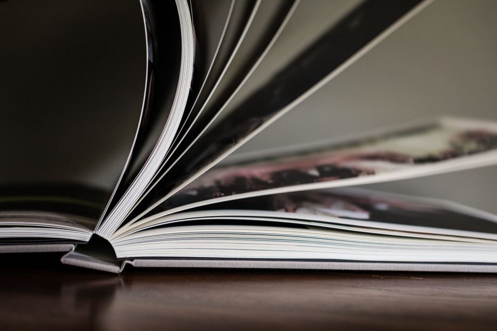 Close-up of an open book with its pages fanned out, resting on a wooden surface. The book appears to contain glossy photographs, and the image focuses on the texture and curve of the pages.