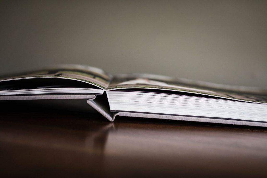 A close-up of an open hardcover book lying flat on a wooden surface, with its pages slightly curved upward and a blurred background.