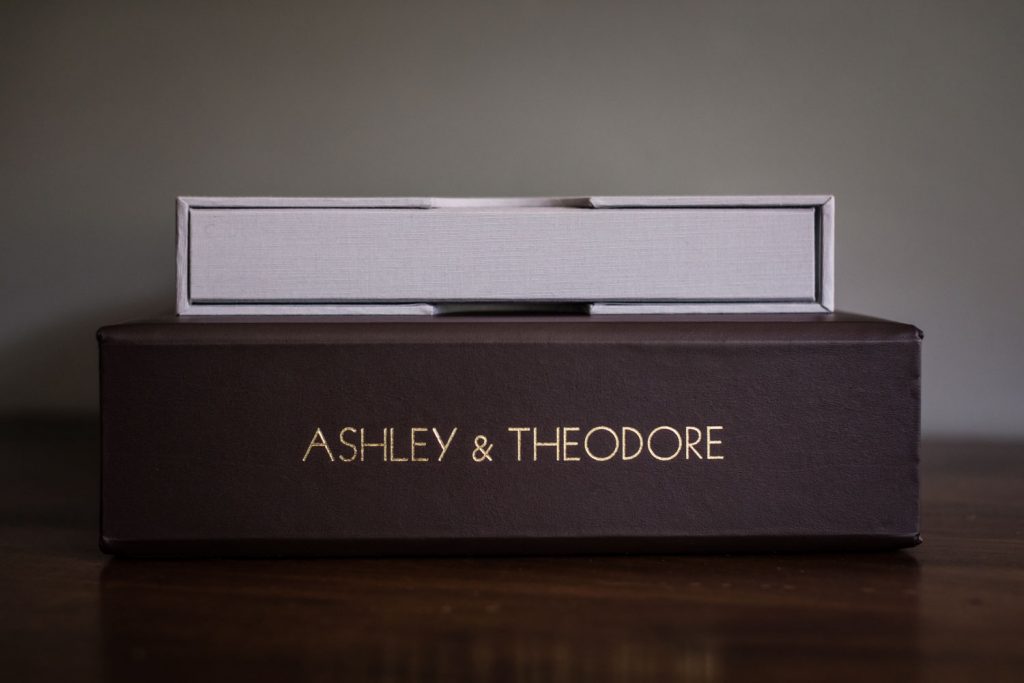 A brown box with gold text reading ASHLEY & THEODORE sits on a wooden surface, with a smaller, light gray box stacked on top.