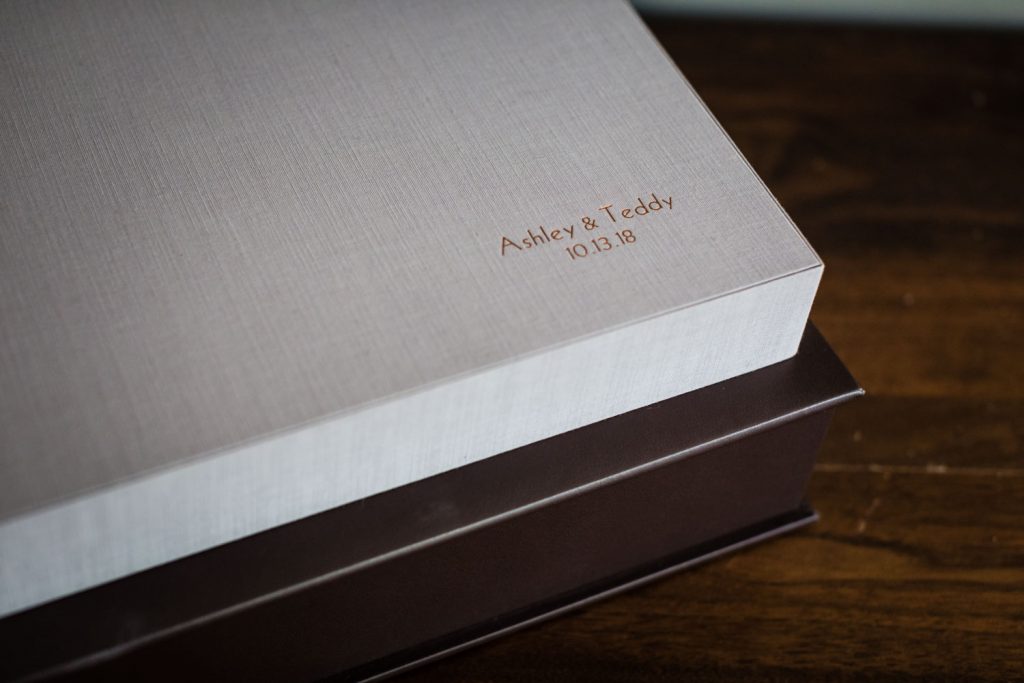 A close-up of a light gray, textured box with Ashley & Teddy 10.13.18 engraved in gold on the lid, resting on top of a dark brown box on a wooden surface.