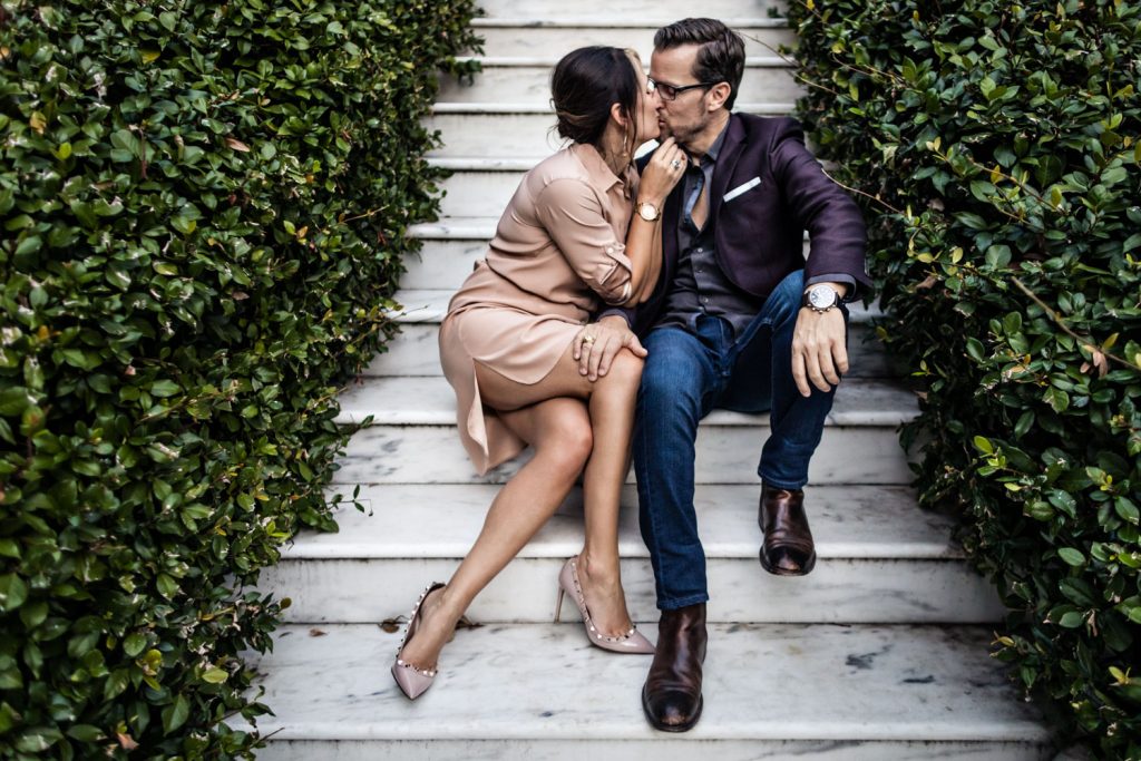 A couple sits on stone steps surrounded by green bushes. The woman wears a light pink dress and a watch, and the man wears a dark jacket, jeans, and boots. They are kissing, and both wear sunglasses.