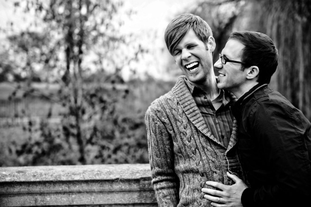 Two people are standing outdoors, laughing together. One is wearing a knit cardigan, and the other has glasses and a dark jacket. They are close, with a blurred background of trees and a stone railing. The image is in black and white.