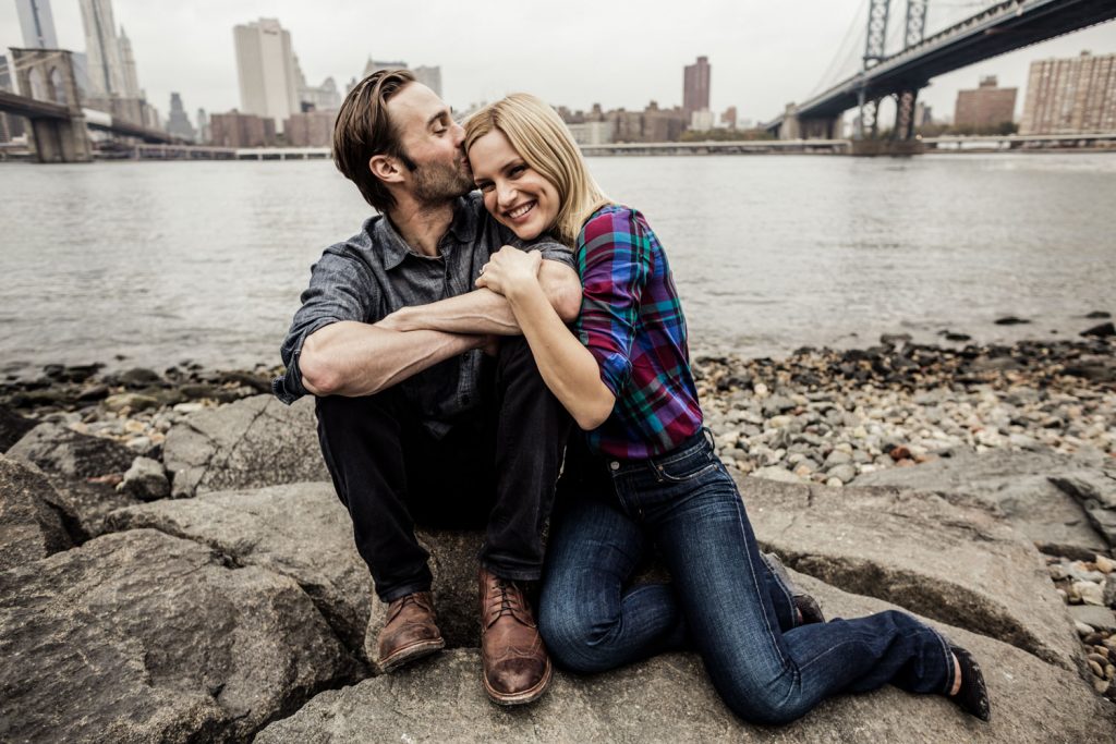 A couple sits on rocks by the water, with a cityscape and bridge in the background. The man kisses the woman's forehead while she smiles, holding his hand. Both are casually dressed, with a cloudy sky overhead.