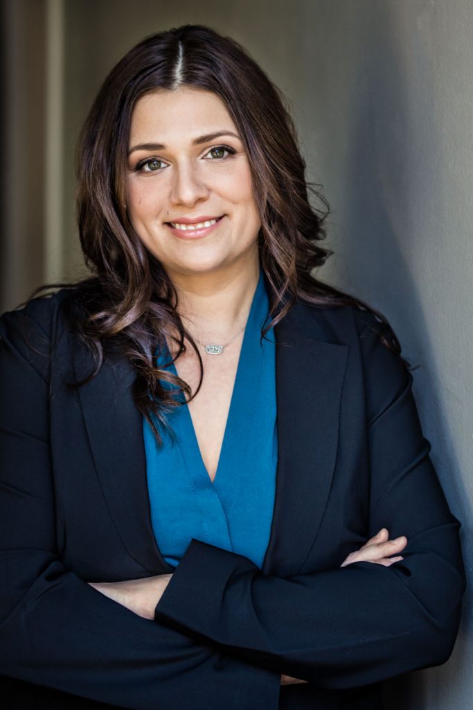 A woman with long dark hair smiles while standing with her arms crossed. She wears a blue blouse and a black blazer, set against a neutral background.