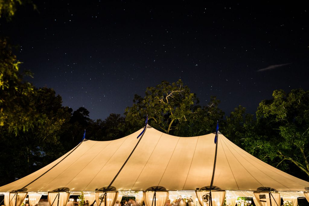 A large, white tent is set up outdoors at night, illuminated warmly from within. The starry sky above and surrounding trees create a serene atmosphere.