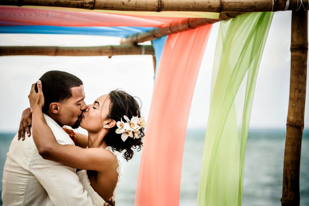 A bride and groom kiss passionately under a bamboo arch adorned with colorful flowing fabrics in orange, green, and blue. The bride has white flowers in her hair, and the ocean is visible in the background.