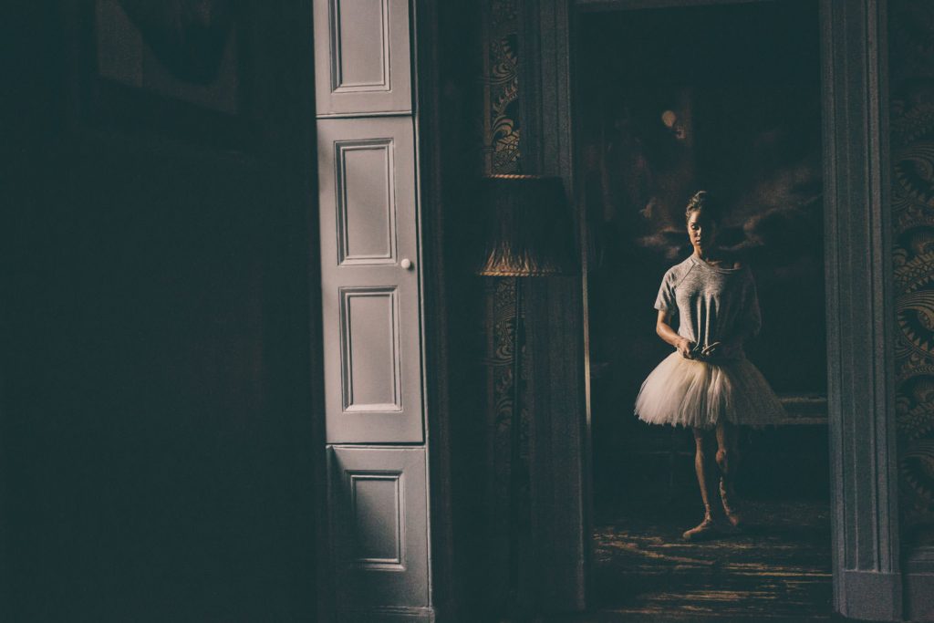 A person wearing a ballet tutu stands in a dimly lit room, captured through an open doorway. The light casts soft shadows, creating a dramatic and mysterious atmosphere.