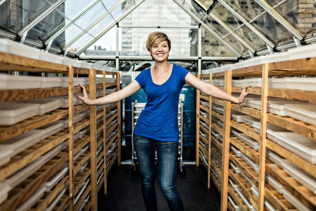 A person with short hair and a blue shirt stands smiling inside a greenhouse, surrounded by wooden shelves with trays. The greenhouse has a transparent roof, allowing natural light to illuminate the space.