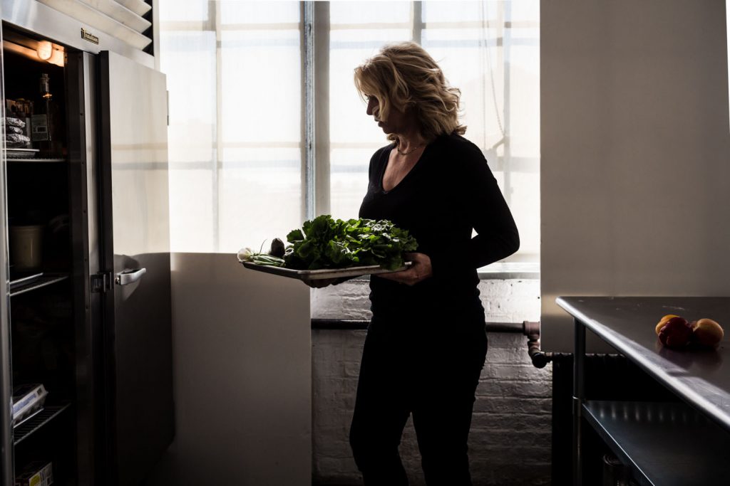 A person in a dimly lit kitchen holds a tray of fresh greens and vegetables. They stand by an open refrigerator, with natural light streaming in through a large window in the background.