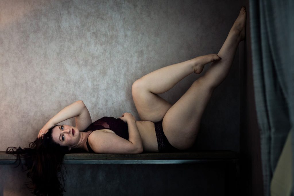 A woman with long dark hair lies on her back on a narrow surface, wearing dark lingerie. She is positioned against a textured wall, her legs raised with one knee bent. The lighting casts soft shadows, and a curtain is partially visible on the right.
