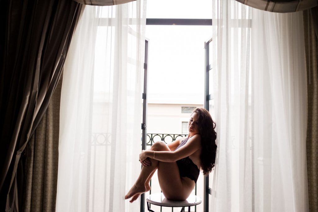 A girl with long hair is sitting on a stool in front of an open window, partially covered by sheer curtains. The room is softly lit by natural light coming through the window, creating a serene atmosphere.