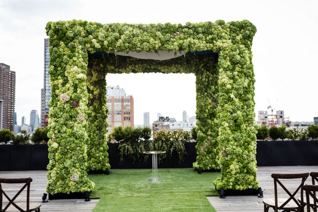 A floral wedding arch made entirely of green and white hydrangeas stands on a rooftop with city buildings in the background, surrounded by greenery and wooden chairs on a grassy aisle.