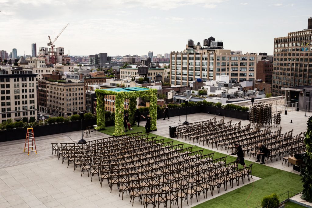 A rooftop venue set up for an outdoor event, with rows of chairs facing a greenery-covered archway, surrounded by city buildings and a few people preparing the space.