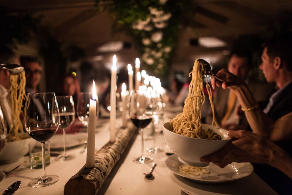 A dimly lit dinner scene features people seated around a table decorated with candles and greenery. A bowl of spaghetti is being served, and glasses of red wine are visible, creating a warm and inviting atmosphere.