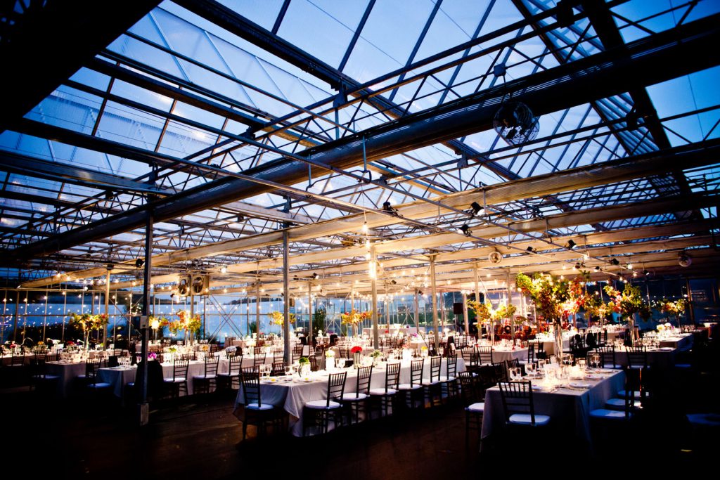 A spacious glass-ceilinged venue arranged for a formal event. Tables are adorned with white cloths, floral centerpieces, and elegant chairs. Soft lighting creates a warm atmosphere, while the evening sky is visible through the glass structure.