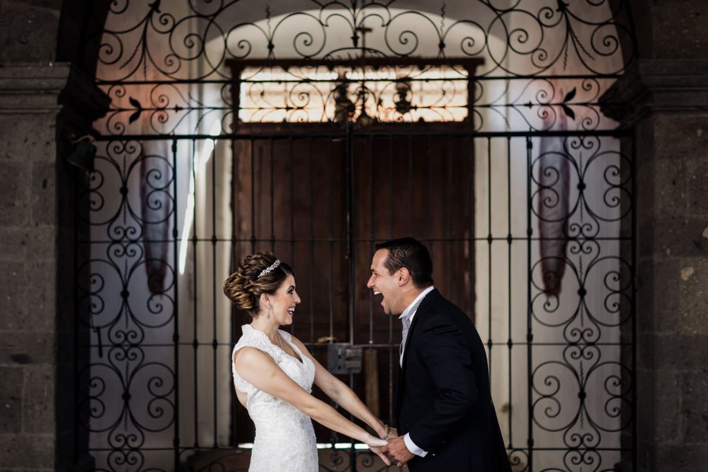 A smiling bride and groom stand holding hands in front of an ornate wrought iron gate. The bride wears a white dress, and the groom is in a black suit, both looking happily at each other.
