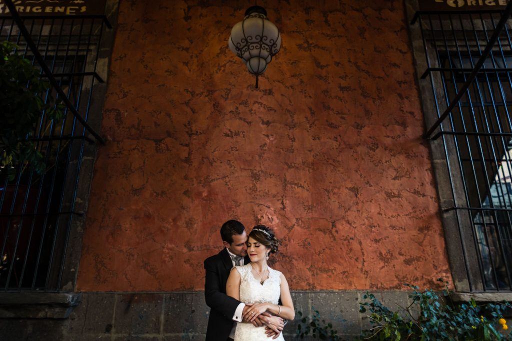 A groom gently embraces a smiling bride in a white dress, standing against a textured reddish-orange wall under a hanging lamp, framed by iron-barred windows and green plants.