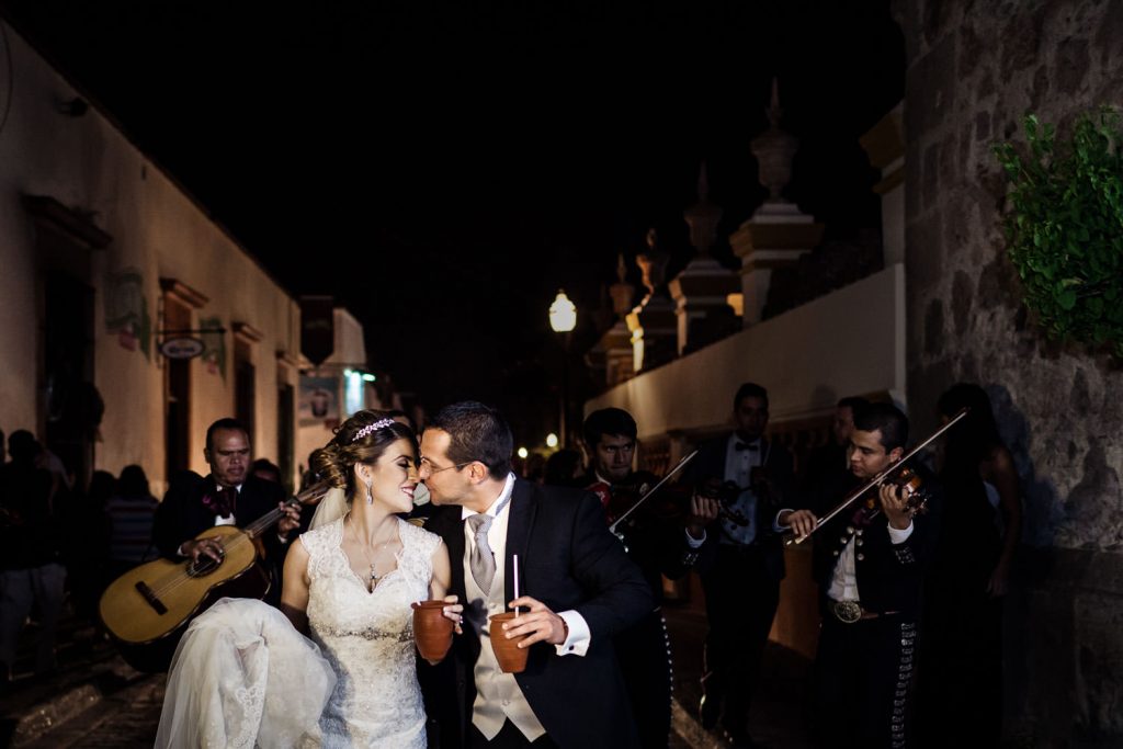 A bride and groom share a kiss while holding cups, surrounded by a mariachi band playing music. Theyre in a dimly lit street, with the bride in a white gown and the groom in a suit.