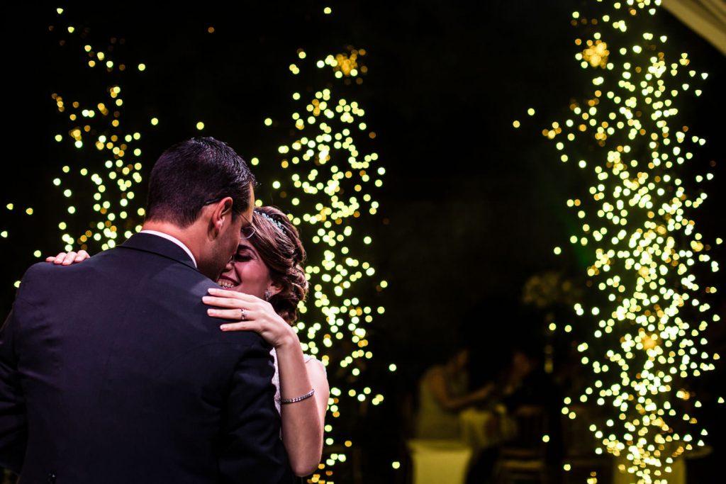 A couple shares a dance at night, smiling and embracing each other, surrounded by warm, glowing string lights and blurred golden bokeh in the background.