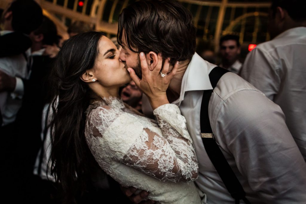 A bride in a lace wedding dress passionately kisses the groom, who wears a white shirt with suspenders, as they embrace on a crowded dance floor.