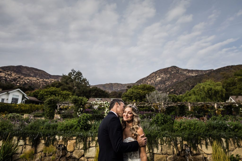 A bride and groom embrace outdoors, smiling with mountains and greenery in the background under a partly cloudy sky. The bride wears a white dress and the groom is in a dark suit. A stone wall and garden plants are visible behind them.