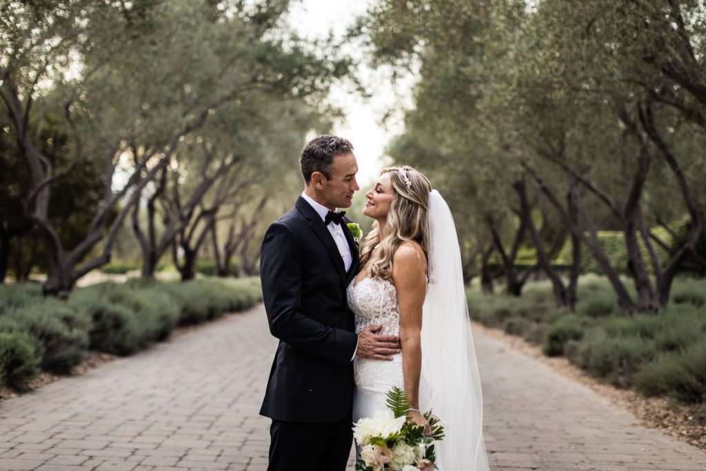 A bride and groom stand on a tree-lined stone pathway, gazing lovingly at each other. The bride wears a white dress with a veil and holds a bouquet, while the groom is in a black tuxedo.