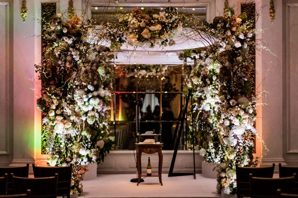 A lavish indoor wedding altar decorated with abundant white and pastel flowers, greenery, and twinkling lights, with a small wooden table and microphone stand in front, surrounded by empty chairs.