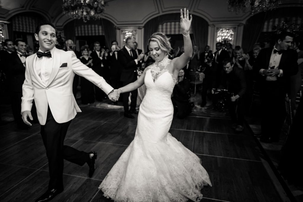 A bride and groom are joyfully dancing, holding hands at a wedding reception. The groom is in a white tuxedo jacket, and the bride is in a lace gown. Guests in formal attire are applauding and smiling in the background.