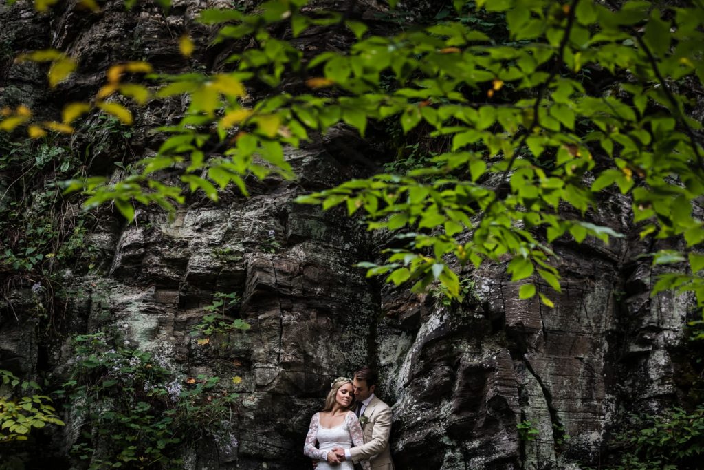 A couple in wedding attire embraces in front of a rocky cliff surrounded by lush greenery, with leafy branches framing the scene above them.