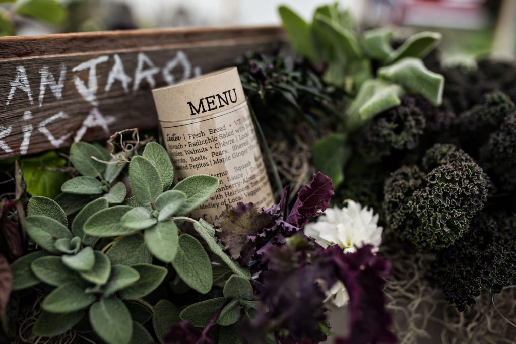 A rolled menu rests among fresh green herbs and leafy vegetables, with a rustic wooden crate partially visible in the background.