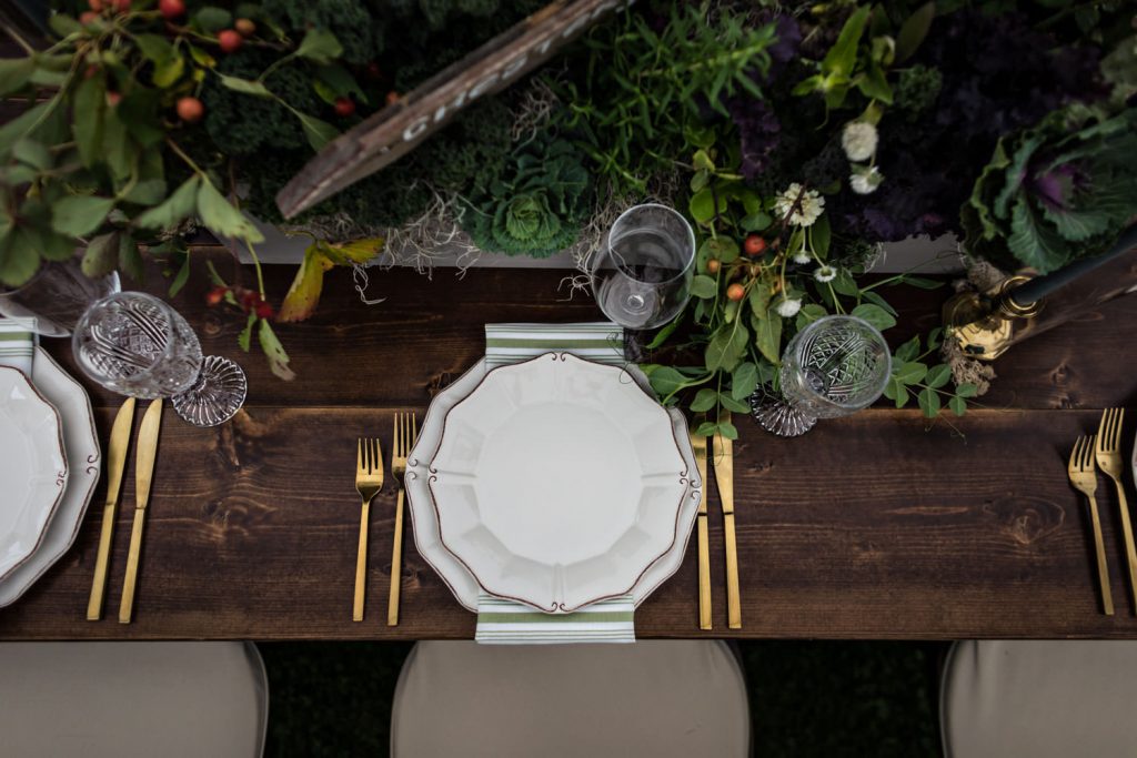 A wooden dining table set with white plates, gold cutlery, crystal glasses, light blue napkins, and decorated with lush greenery and small fruits as a centerpiece.