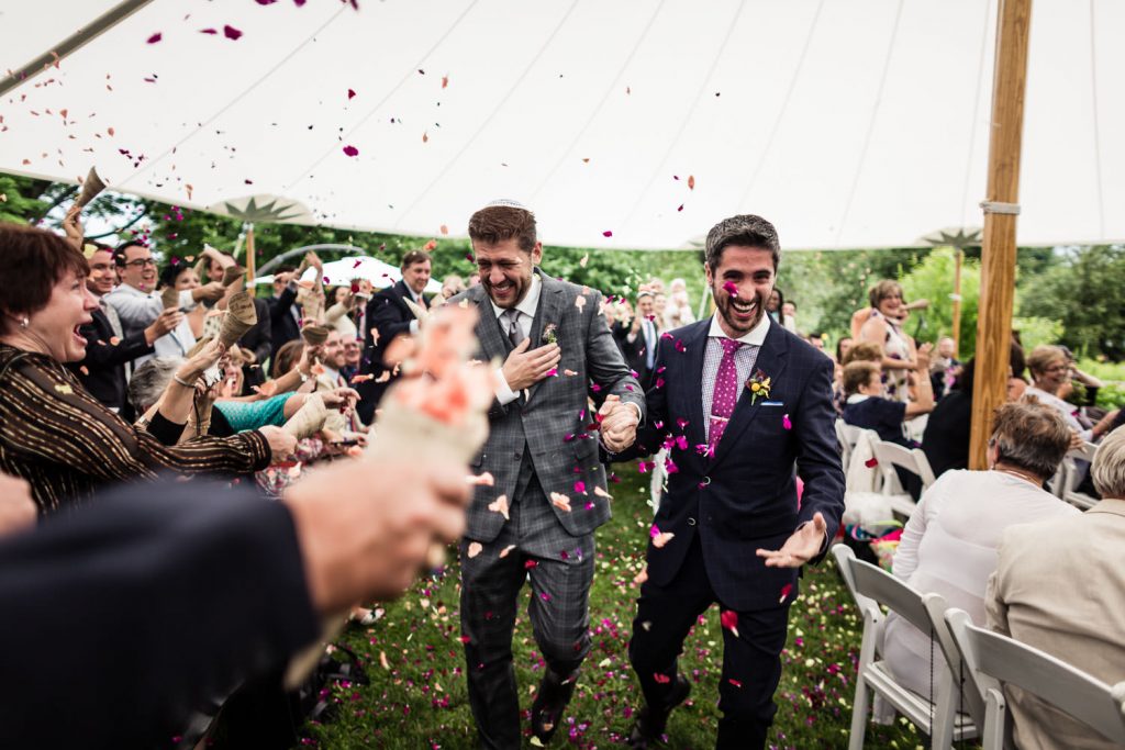 Two grooms joyfully walk down the aisle under a large white tent, surrounded by guests throwing colorful flower petals. They are dressed in suits, one in gray plaid and the other in navy. The seated guests appear happy and celebratory.