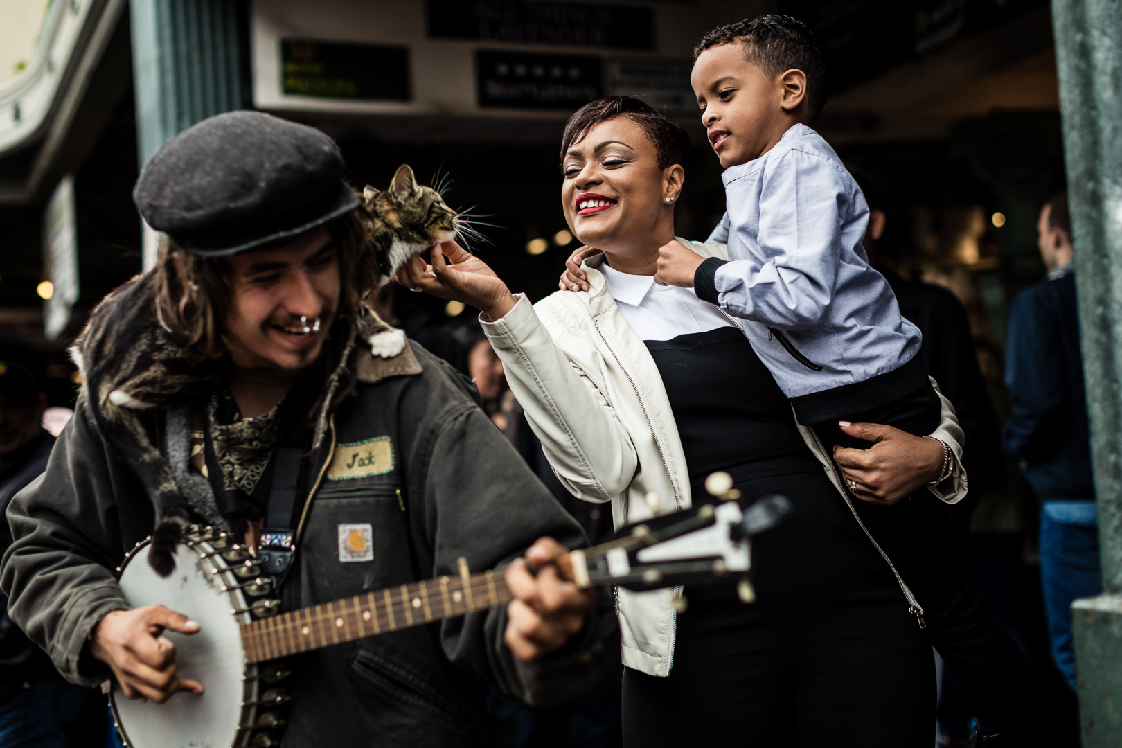 A street musician plays the banjo while a woman holding a child smiles and reaches out to pet a cat resting on the musicians shoulder. The scene captures an urban setting with people visible in the background.