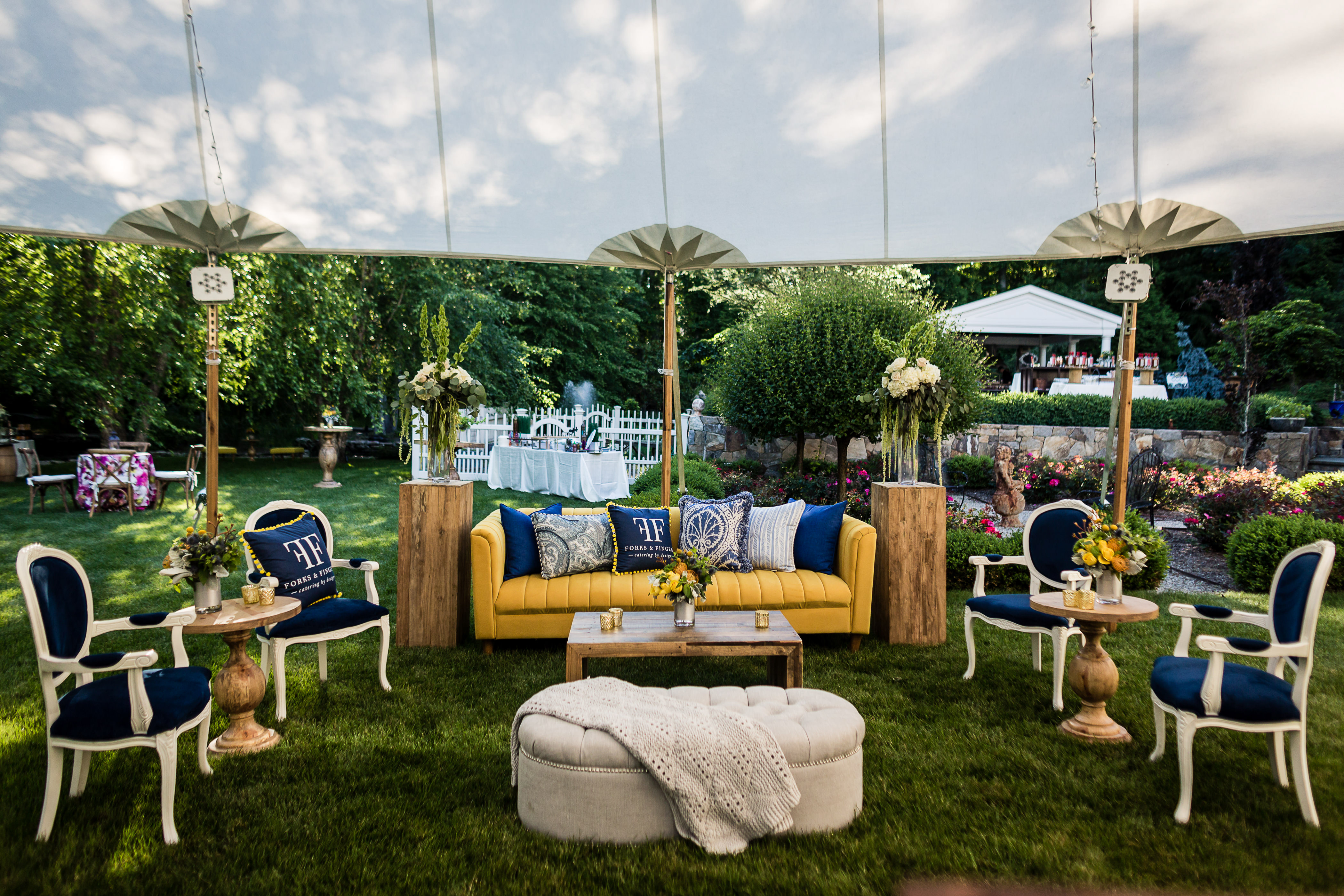 Forks + Fingers Launch Party A stylish outdoor seating area with a yellow sofa, blue cushions, and elegant wooden tables under a white canopy, perfect for a corporate photographer's shoot. Surrounded by lush greenery and floral arrangements, it includes white and blue chairs and a cushioned ottoman on the grass.