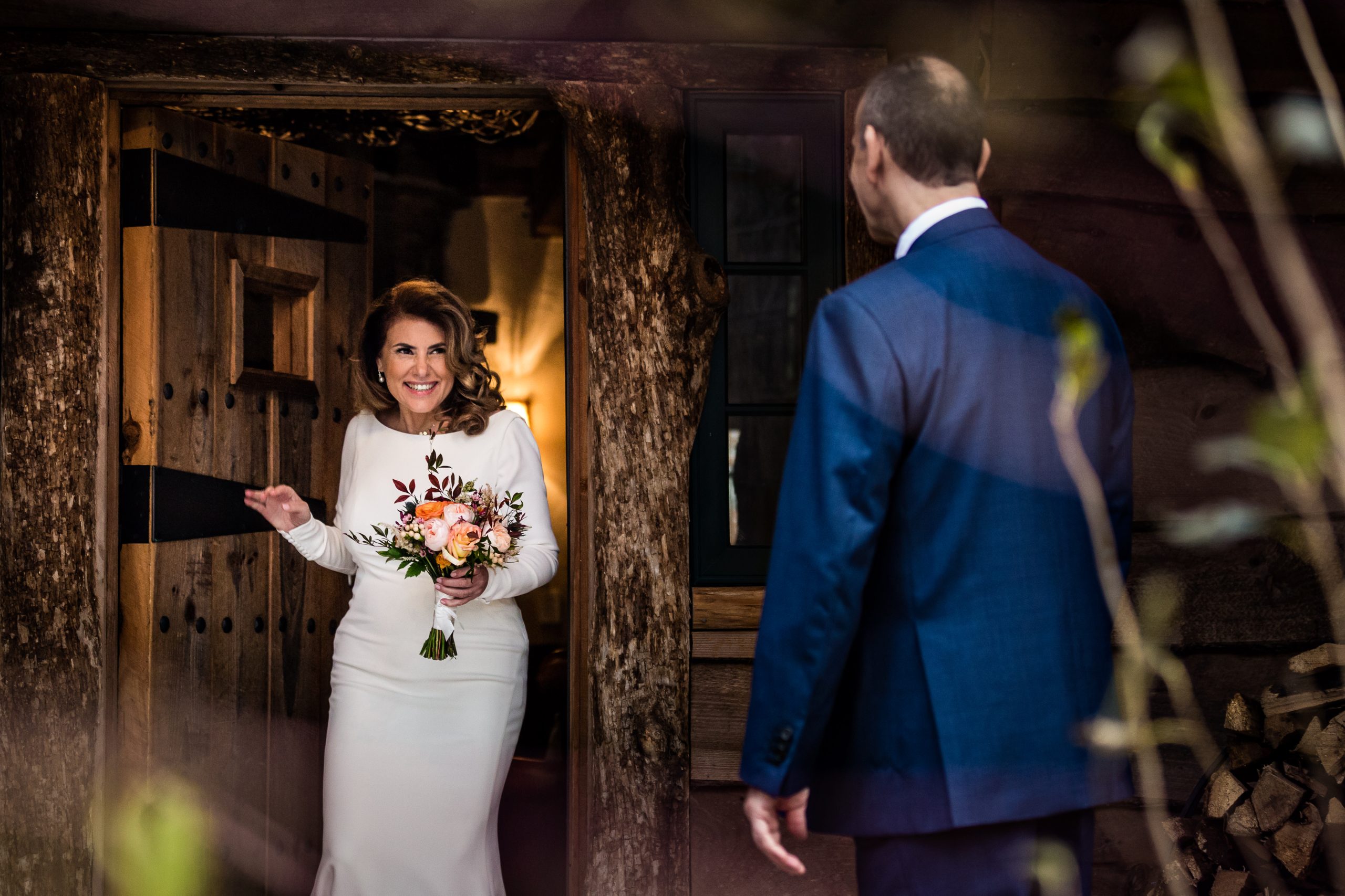 A bride in a white dress holds a bouquet, smiling warmly in a rustic wooden doorway. Her partner in a blue suit faces her, partially turned away. Foliage frames the intimate scene captured by an elopement photographer, creating a romantic atmosphere.