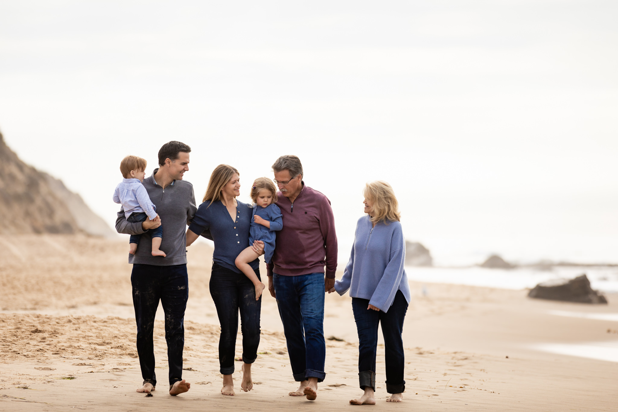 A family walks barefoot on a sandy beach. Two adults hold young children, while an older couple holds hands. They are dressed in casual clothes, and the ocean is visible in the background on a cloudy day.