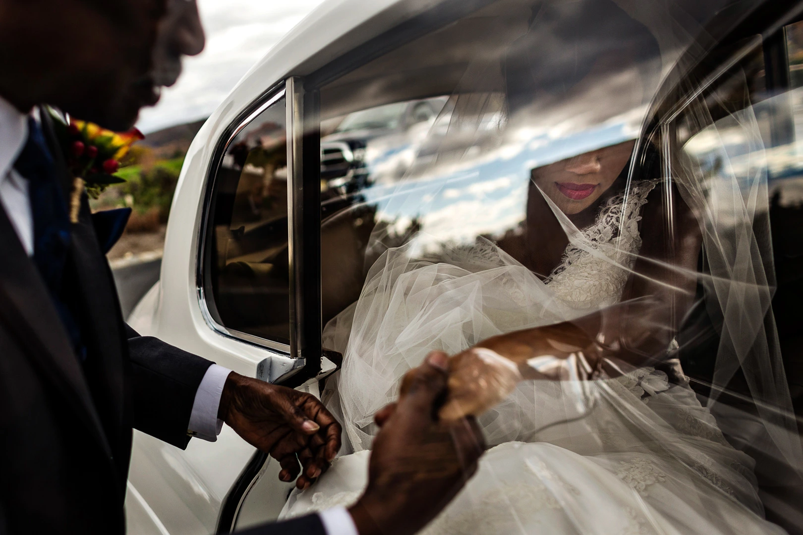 A bride in a white dress sits inside a car, partially visible through the window. Her veil covers her face, and a person in a suit helps her with the gown. The reflection of the sky and clouds is seen on the car's surface.