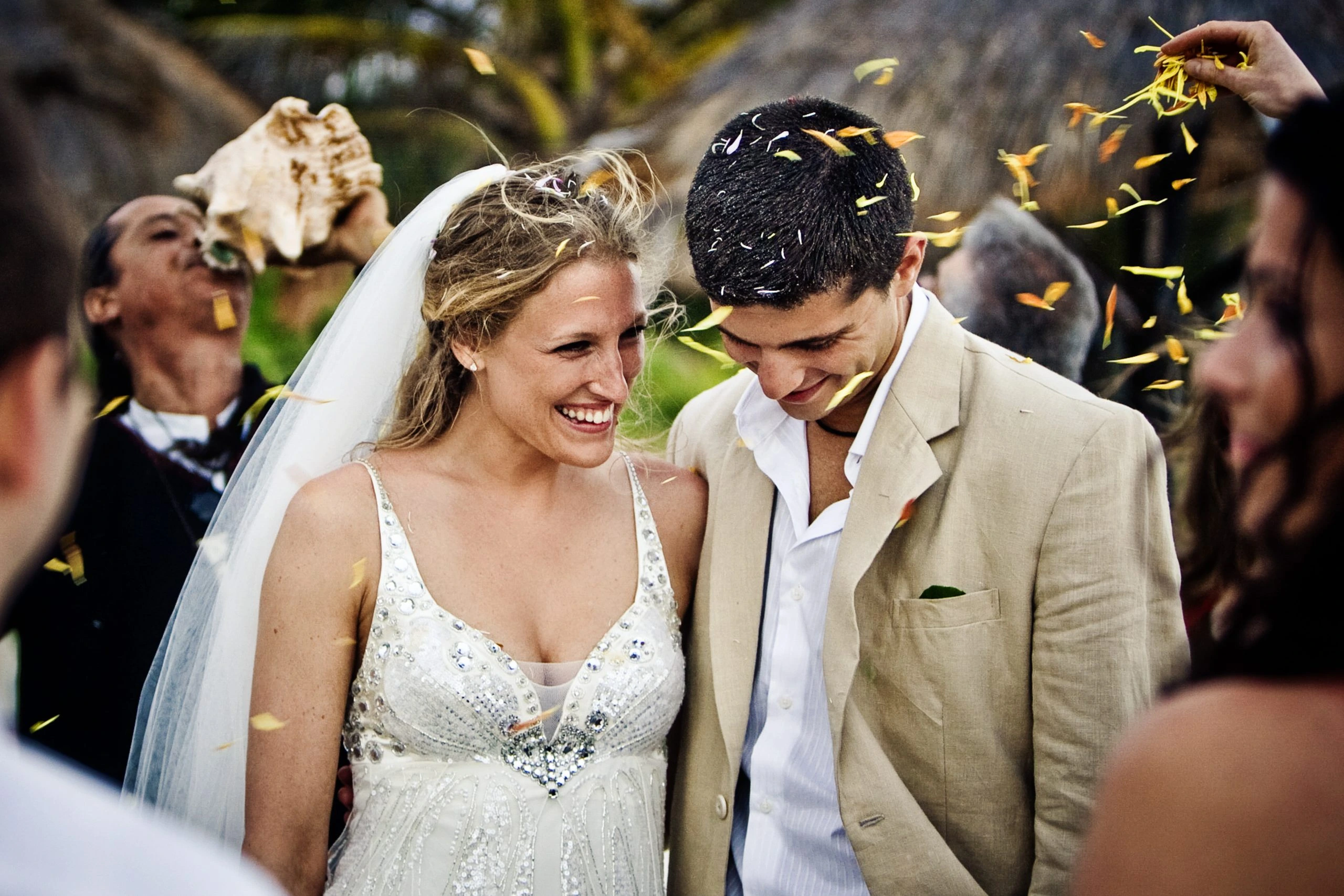 A bride and groom smile joyfully as flower petals are tossed in celebration. The bride wears a sparkling white dress and veil, while the groom is in a beige suit. A man in the background blows into a conch shell. The scene is outdoors.
