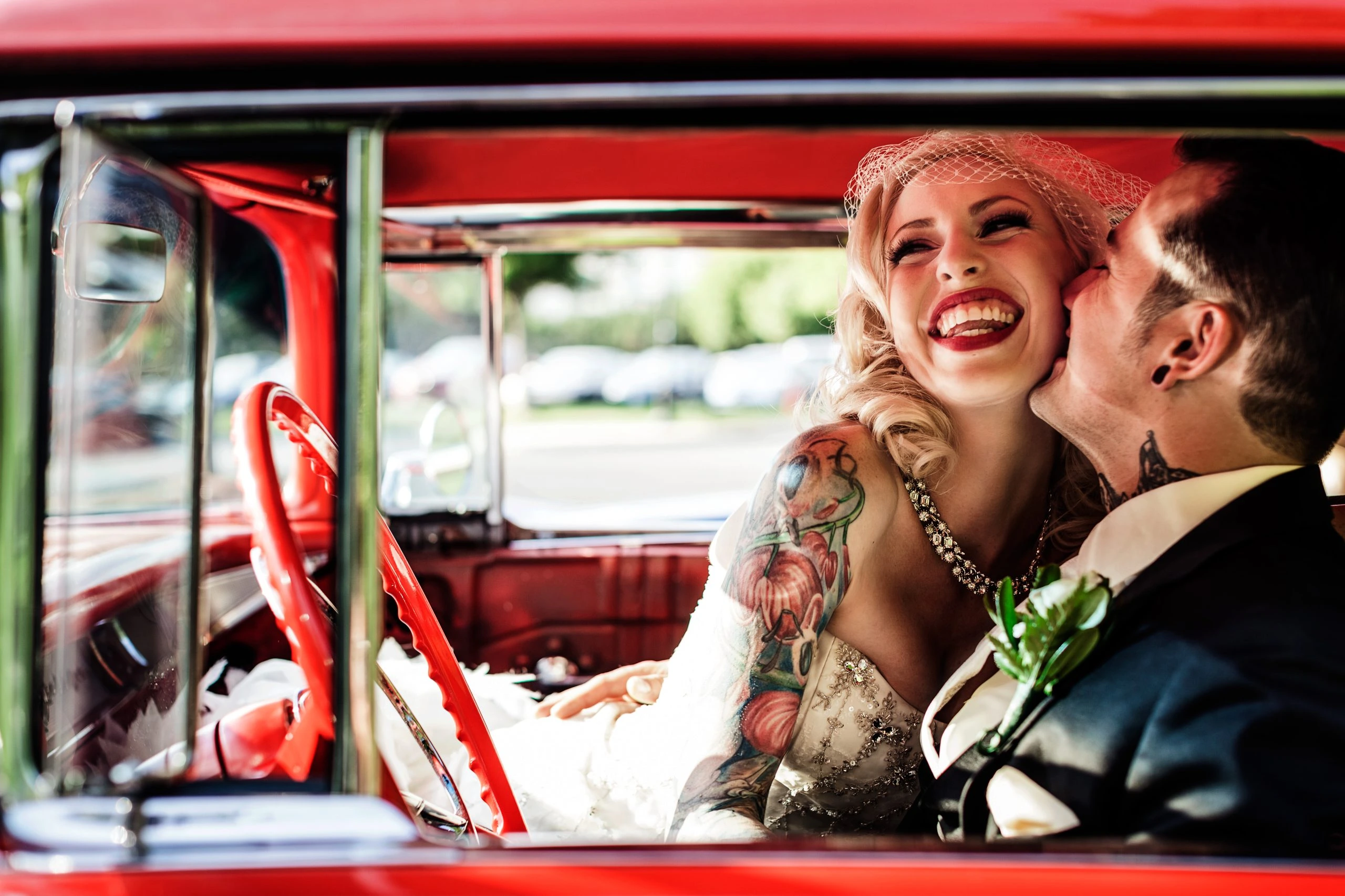 A joyful couple sits in a vintage red car. The woman, wearing a white dress and veil, laughs heartily, while the man, in a dark suit and adorned with tattoos, smiles affectionately beside her.