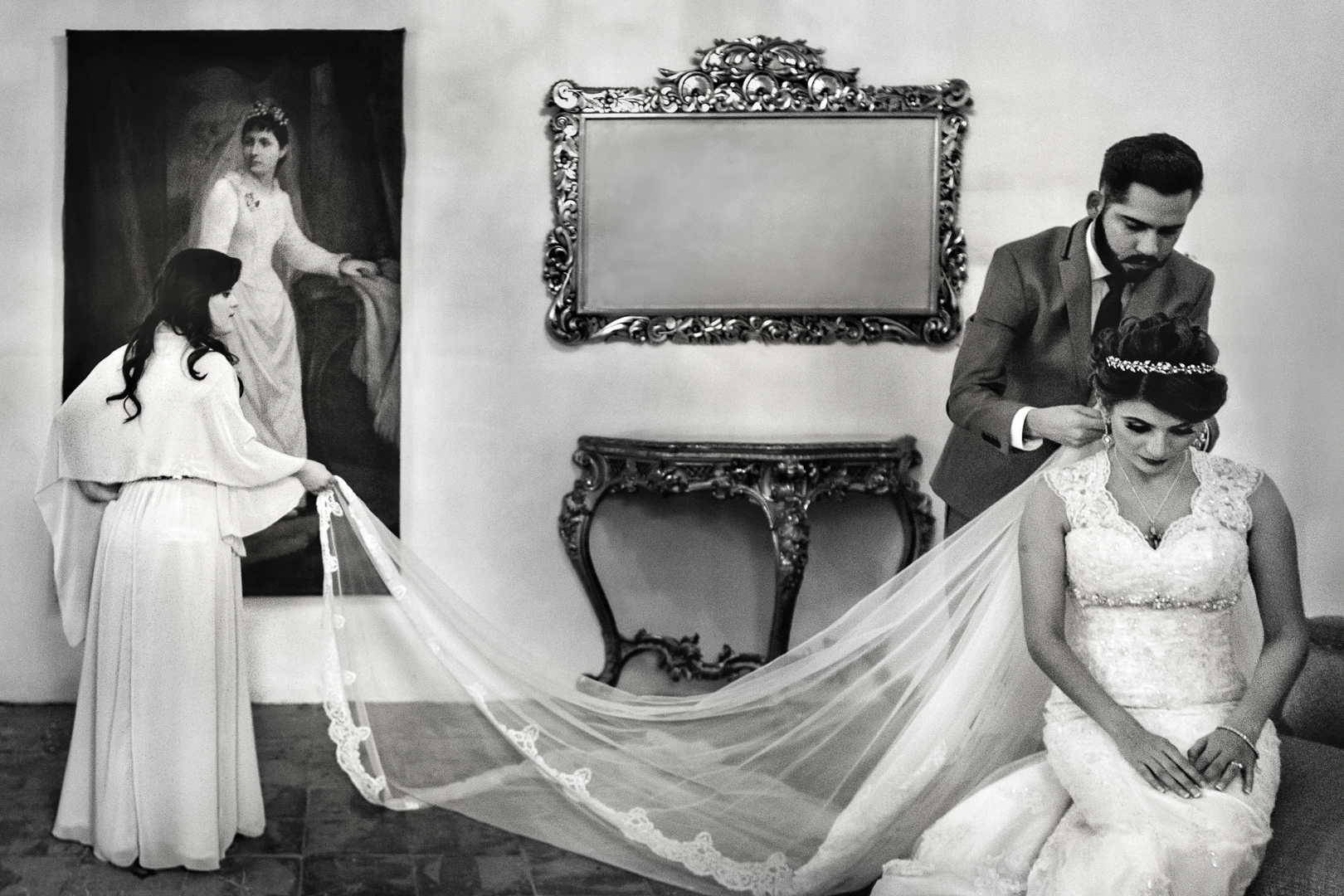 A black and white photo of a bride in a lace gown and tiara, seated, with a male adjusting her veil. Another female holds the end of the veil. They are in a vintage room with an ornate mirror and a portrait on the wall.